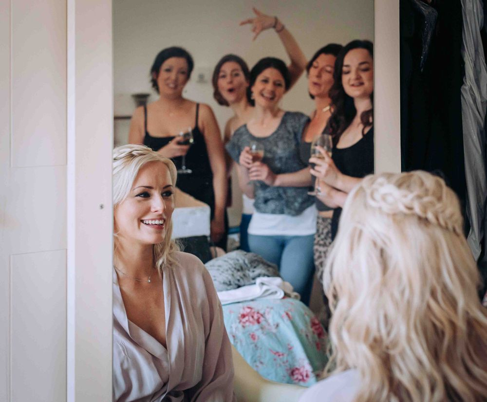 A bride smiles at her waving bridesmaids in the mirror of her dressing room