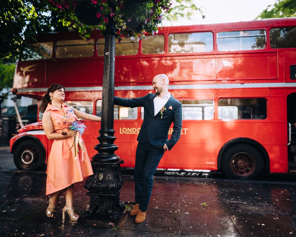 A woman in a peach wedding dress holds onto an ornate black lamppost in front of a Red London bus