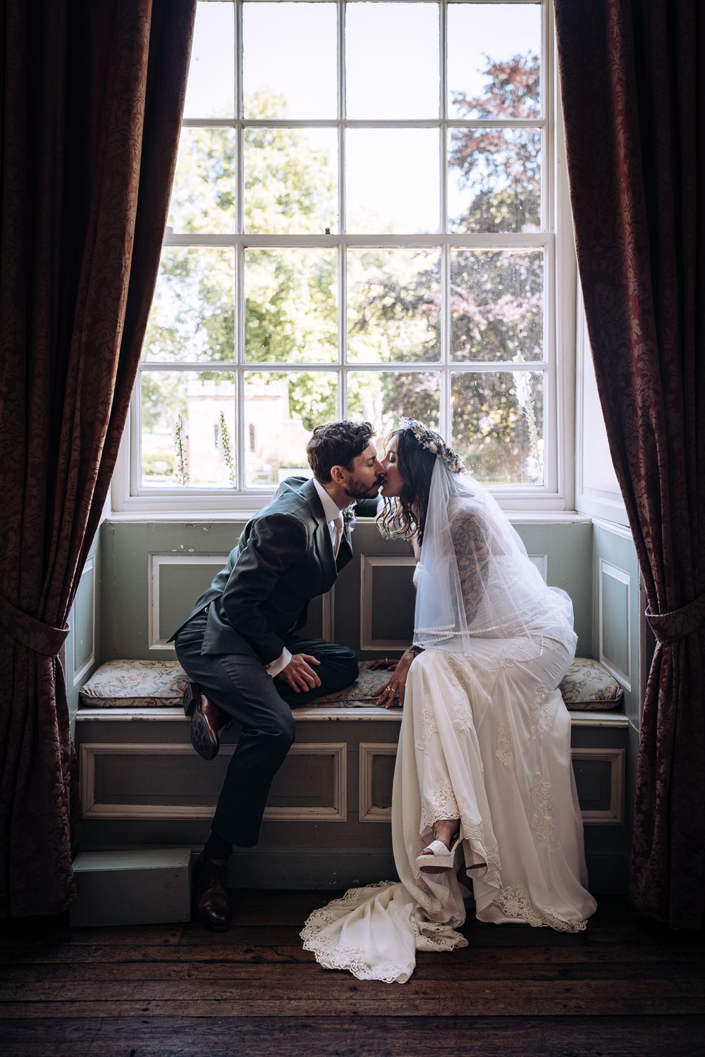 A groom in smart morning suit and a tattooed woman in a wedding dress sit in a large bay window at Hartlebury Castle