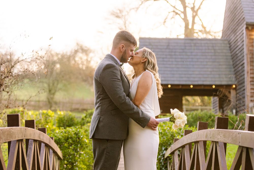 A bride a groom embrace and share a tender kiss stood on a small and ornate wooden bridge