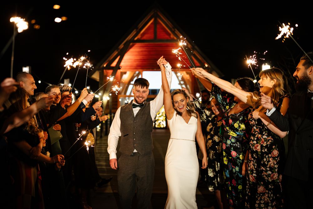 A bride and groom hold their hands together in the air between guests who are holding up lit sparklers