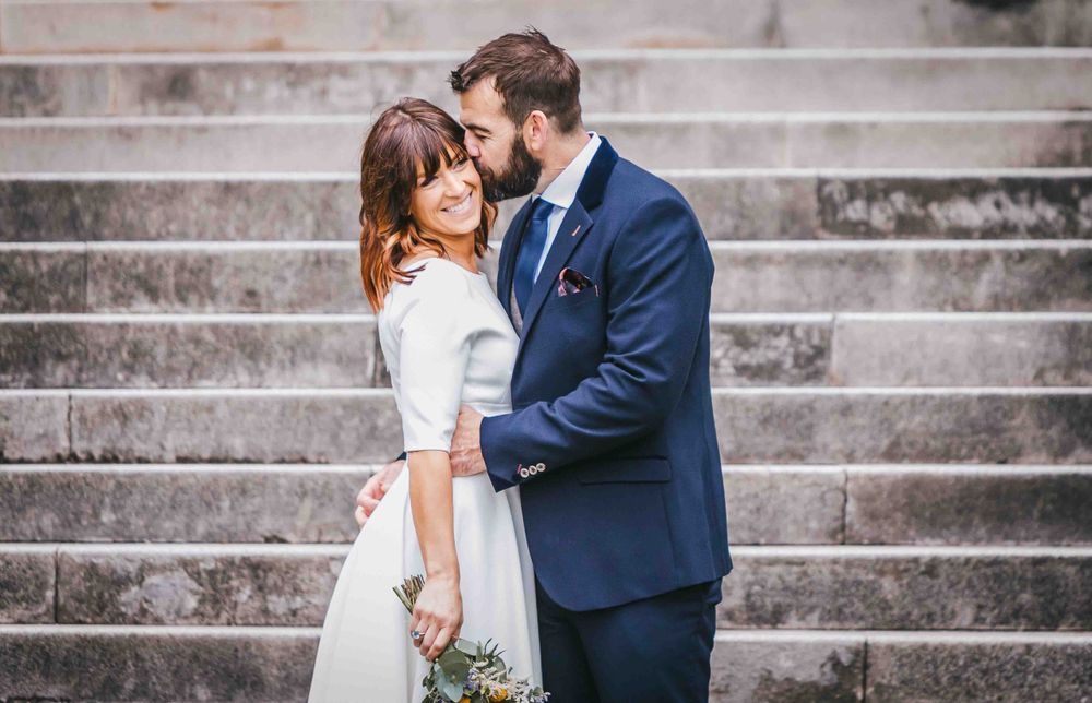 A groom kisses his new brides at the foot of some ornate steps as she smiles at the camera