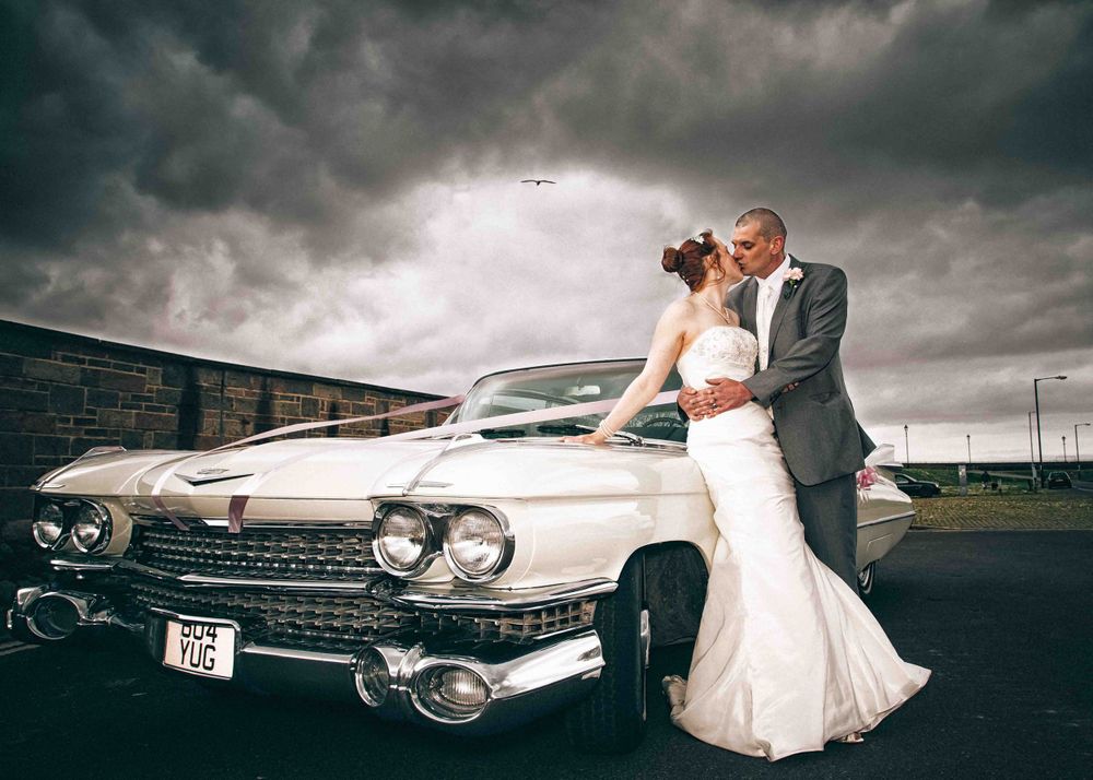 A Bride and Groom kiss against next to a beautiful white Cadillac underneath a moody contrasting sky